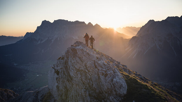 Zwei Wanderer stehen mit Wanderstöcken auf einem felsigen Gipfel bei Sonnenaufgang, Bergpanorama im Hintergrund.