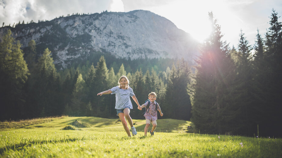 Zwei Kinder laufen lachend über eine grüne Wiese vor dichtem Wald und Bergkulisse im Sonnenlicht.