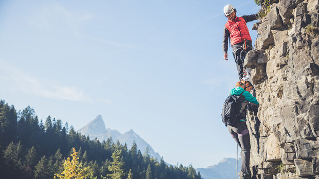 Zwei Personen mit Kletterausrüstung steigen gesichert an einer Felswand, im Hintergrund bewaldete Berge unter blauem Himmel.