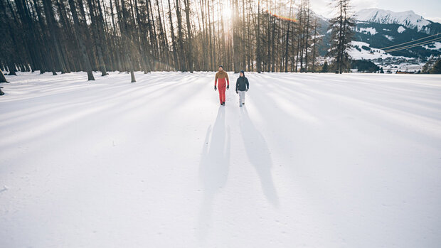 Zwei Personen gehen über eine schneebedeckte Wiese mit langen Schatten, im Hintergrund Wald und Tal.