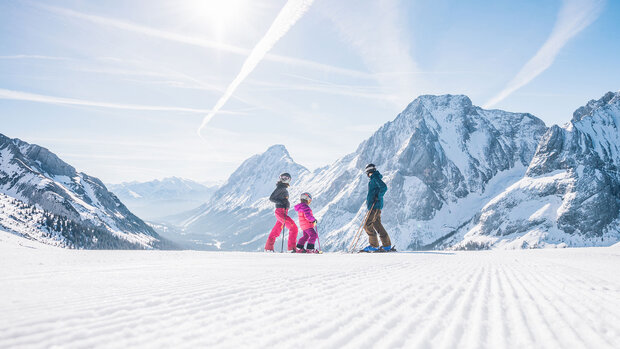 Familie mit Skiern steht auf einer präparierten Piste und blickt auf ein weites, schneebedecktes Bergpanorama.