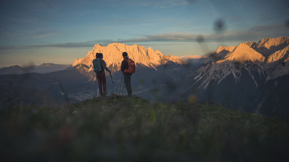 Zwei Wanderer stehen auf einer Anhöhe und blicken auf ein beleuchtetes Bergmassiv im Abendlicht.