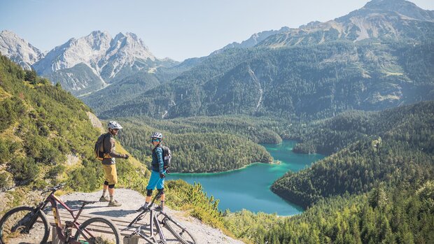 Zwei Mountainbiker stehen mit ihren Fahrrädern auf einem Aussichtspunkt über einem türkisfarbenen Bergsee, umgeben von bewaldeten Bergen.